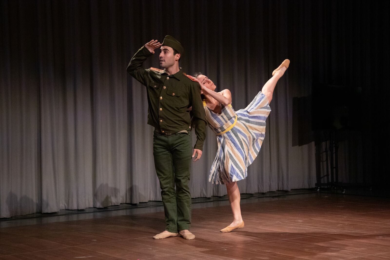 A man in a military uniform salutes while a woman in a striped dress dances behind him in an arabesque in a theatrical setting with a gray curtain behind them.