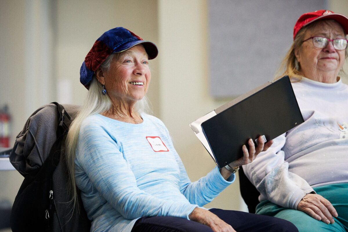 Two women wearing hats are seated while looking off to the right side. The woman on the left is holding an open songbook in her left hand.