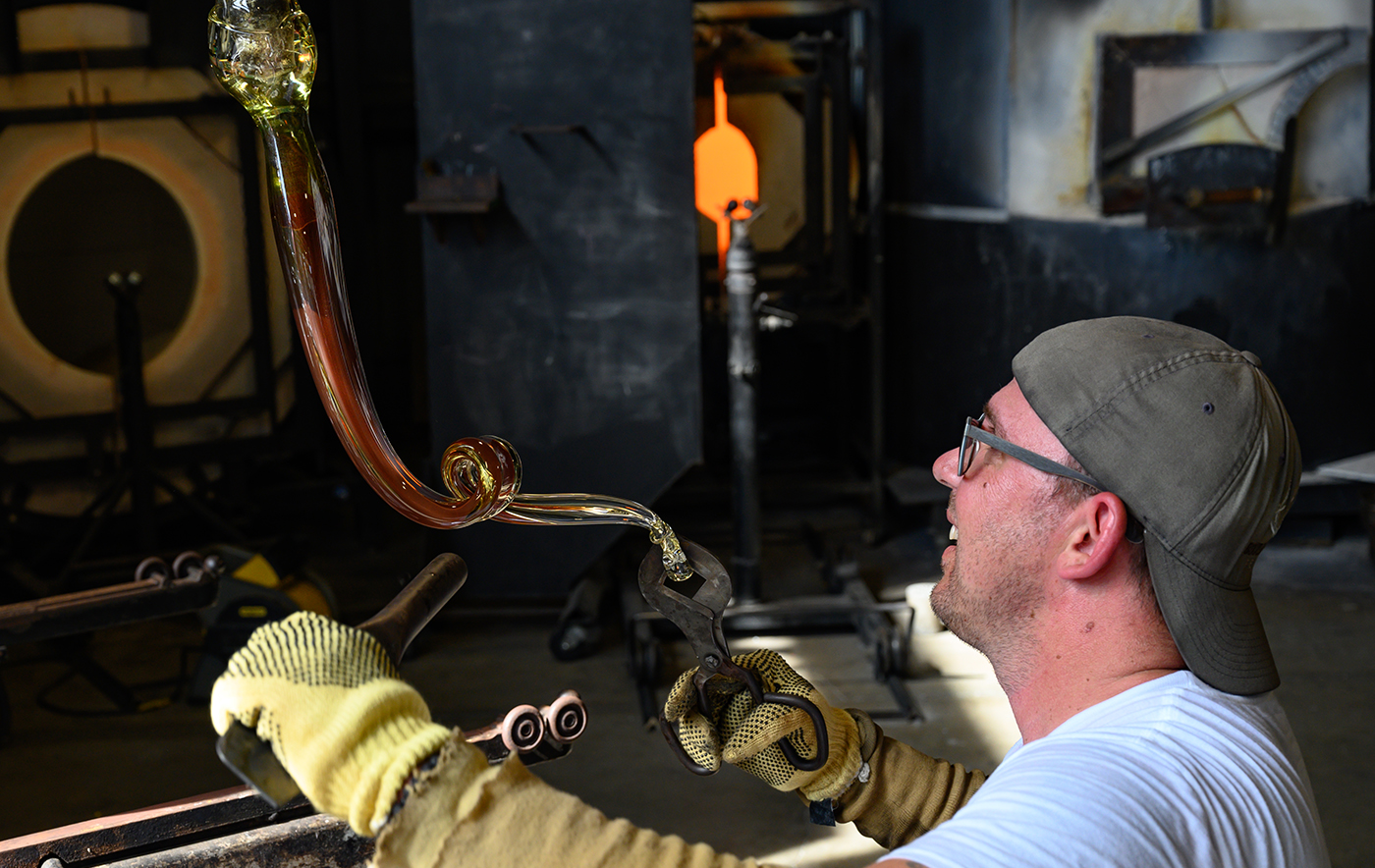 Side profile of a white man wearing protective gloves using tools to hold a glass-blown object. A glass-blowing studio is in the background.