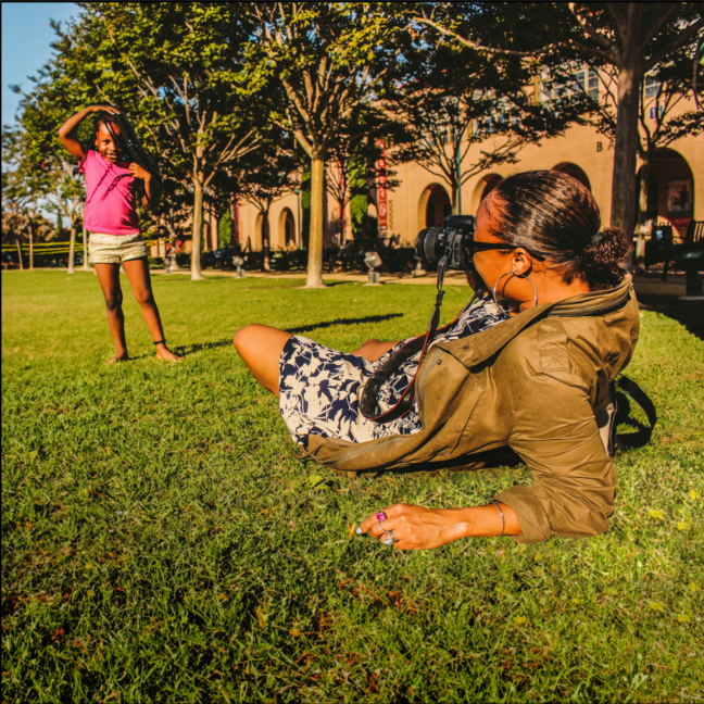 A Black woman sits on the grass leaning back while she takes a photo of her daughter standing in a field with one arm overhead.