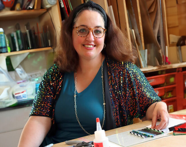 A white woman with shoulder-length brown hair, wearing glasses, is smiling and seated in an art studio. Colorful art materials and tools are on the table in front of her.