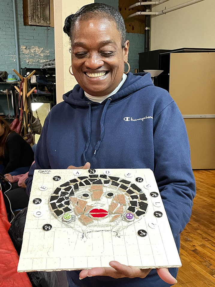 A black woman with her hair pulled back stands smiling while looking down at mosaic art she is holding.