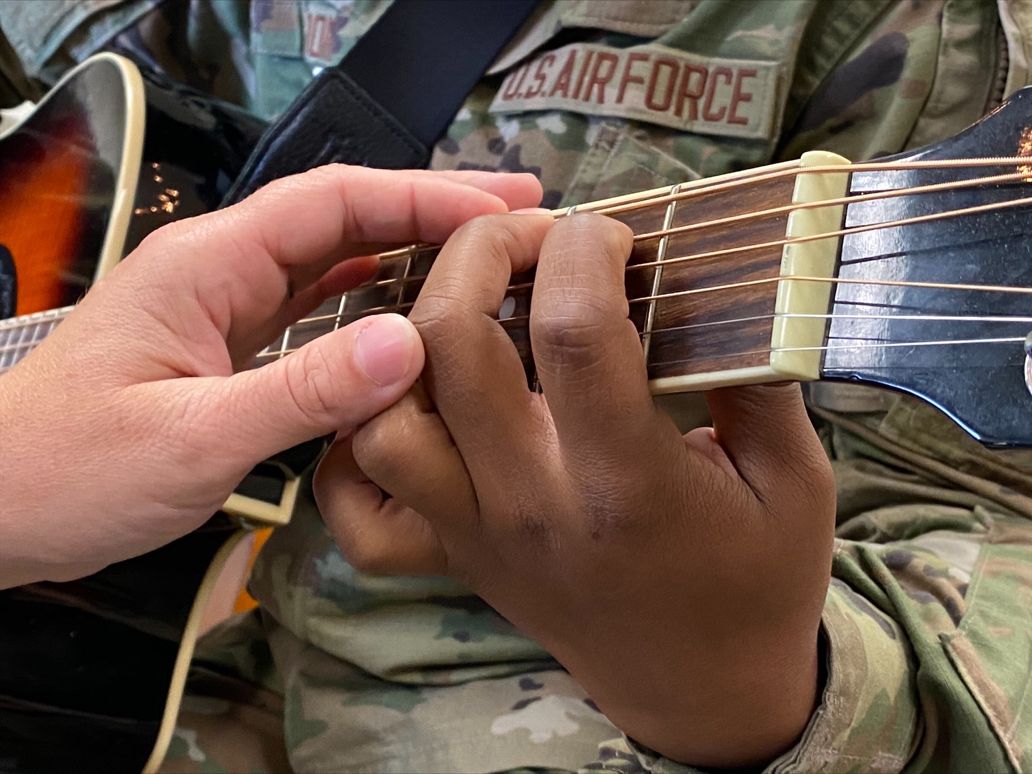 Person in U.S. Air Force uniform with one hand on a guitar neck while a second person's hand touches the guitar and the first person's hand.