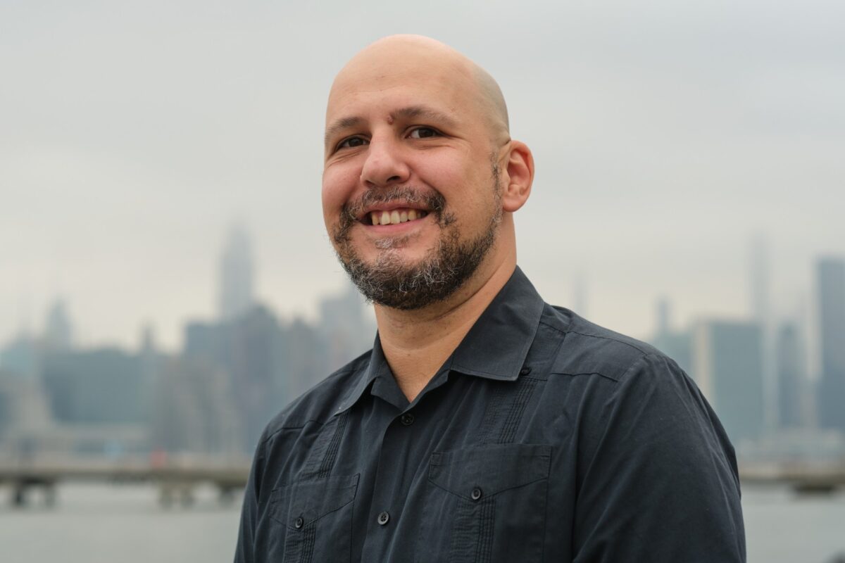 Man with a goatee and dark button down shirt is smiling at the camera with a blurry city skyline in the background.