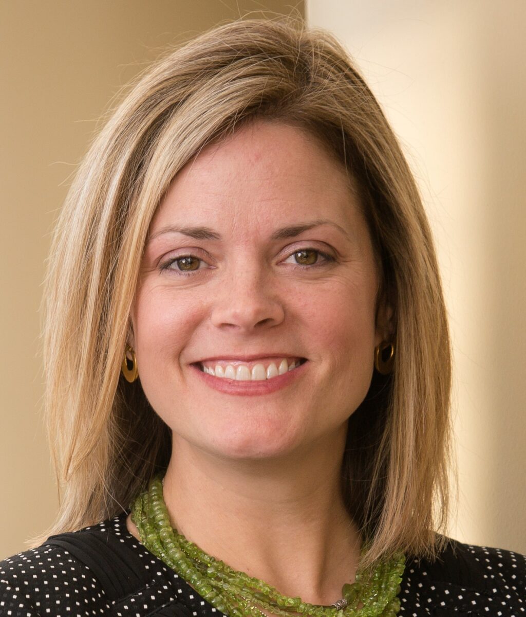 Portrait of a smiling woman with shoulder-length blonde hair, wearing a black and white polka dot top and a green necklace.