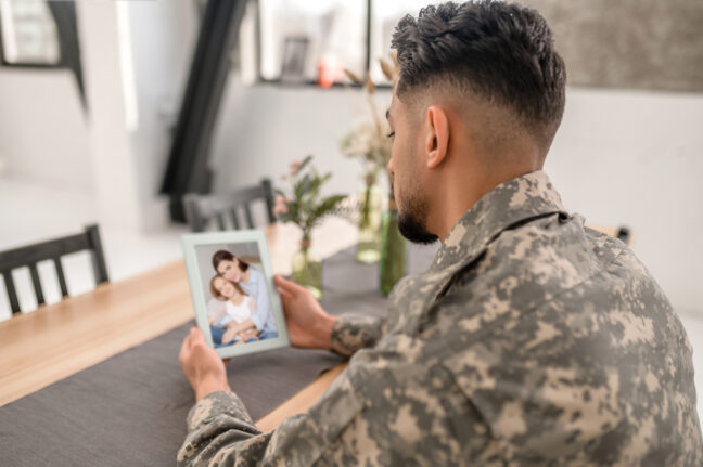 Side view of a man in a military uniform seated at a table and looking at a photo he is holding of a woman and child.