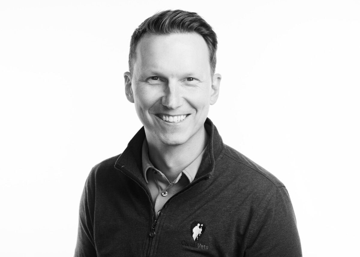 A black and white portrait of a smiling man wearing a 1/4 zip shirt with a CreatiVets logo. 