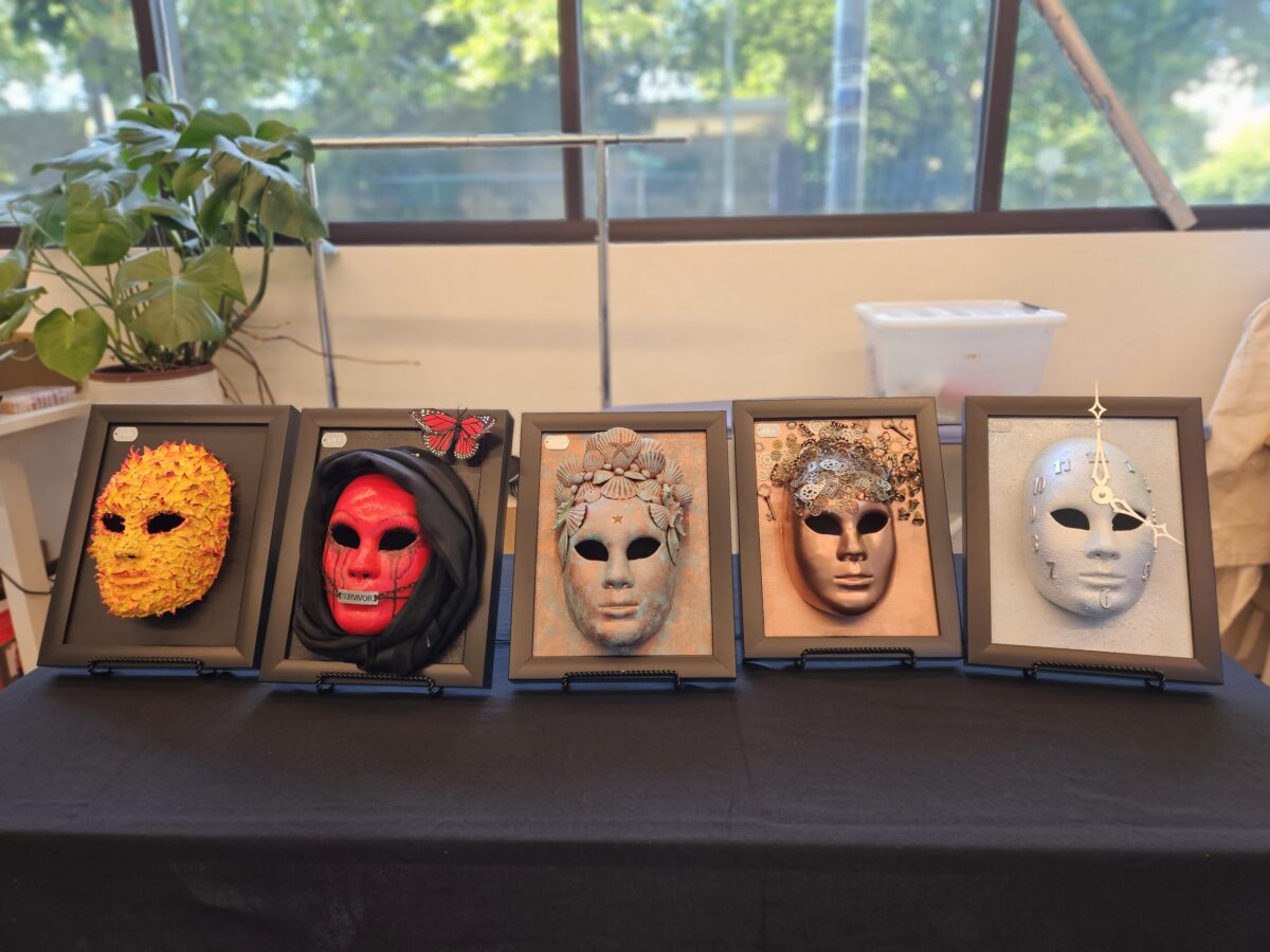 Five colorful, textured masks are arranged on stands on a table. From left to right, the masks feature themes of flora, a hooded red face, delicate floral designs, earthy textures, and a clock motif. 