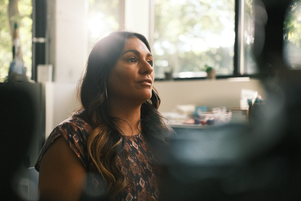 Latina woman with long wavy brown hair and large hoop earrings looking off to the right. She is wearing a patterned top, and is sitting in a sunny room with large windows.