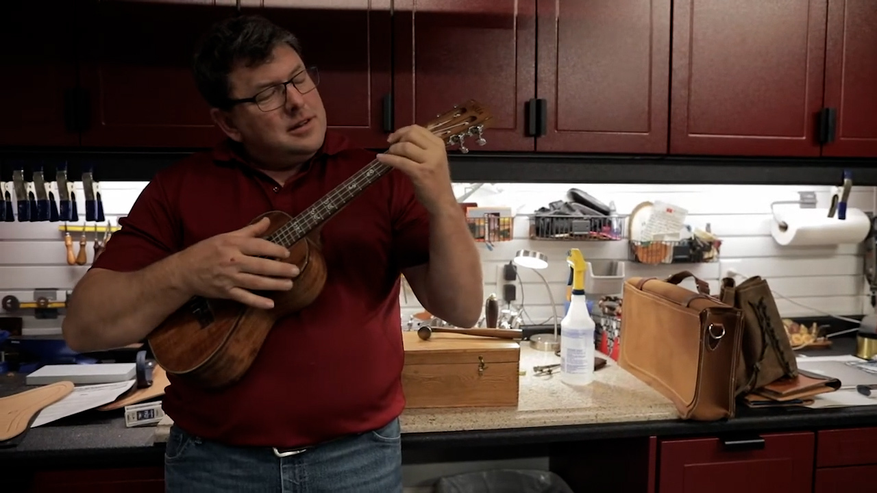 Middle-aged white man strumming a ukulele in a garage workshop space.