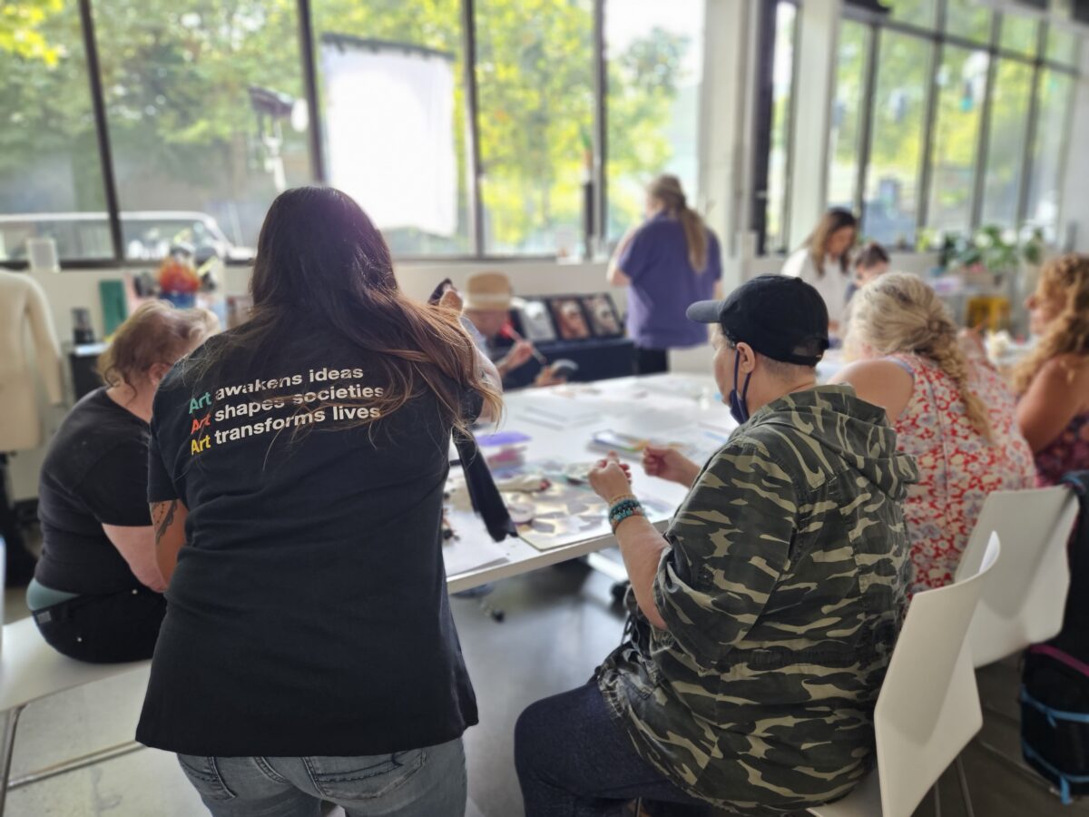 People seated around a large white table engaged in painting and crafts inside a bright room with huge windows. A person in the foreground wears a shirt with inspirational text about art.