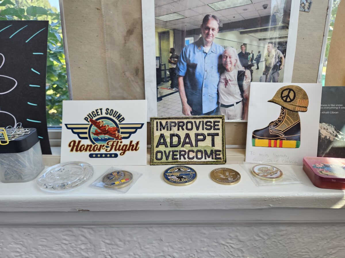 A collection of military memorabilia, patches, and medals sits on a white shelf. One patch reads Puget Sound Honor Flight, another says Improvise Adapt Overcome. A framed photo of two people is in the background.