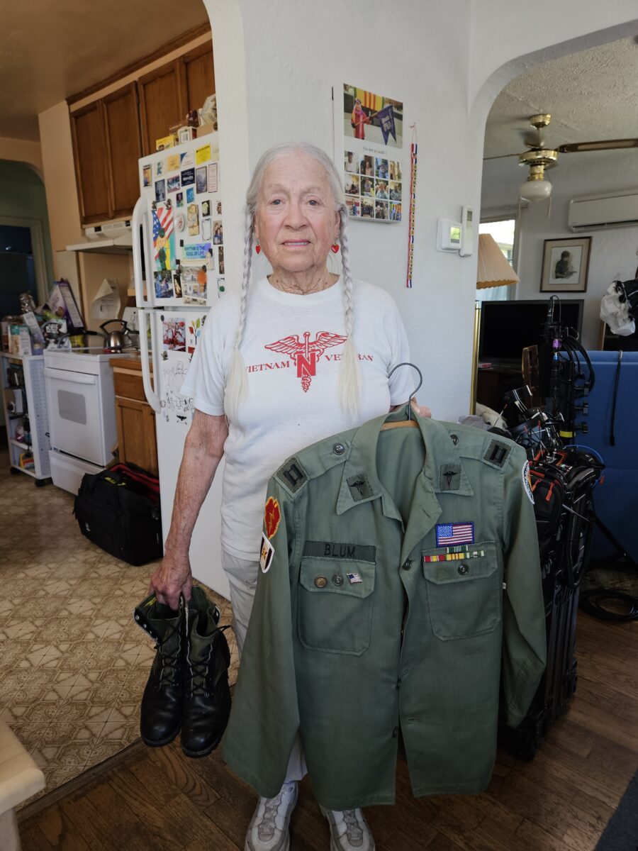 A white woman with long gray hair in two braids holds her US Army uniform and combat boots.