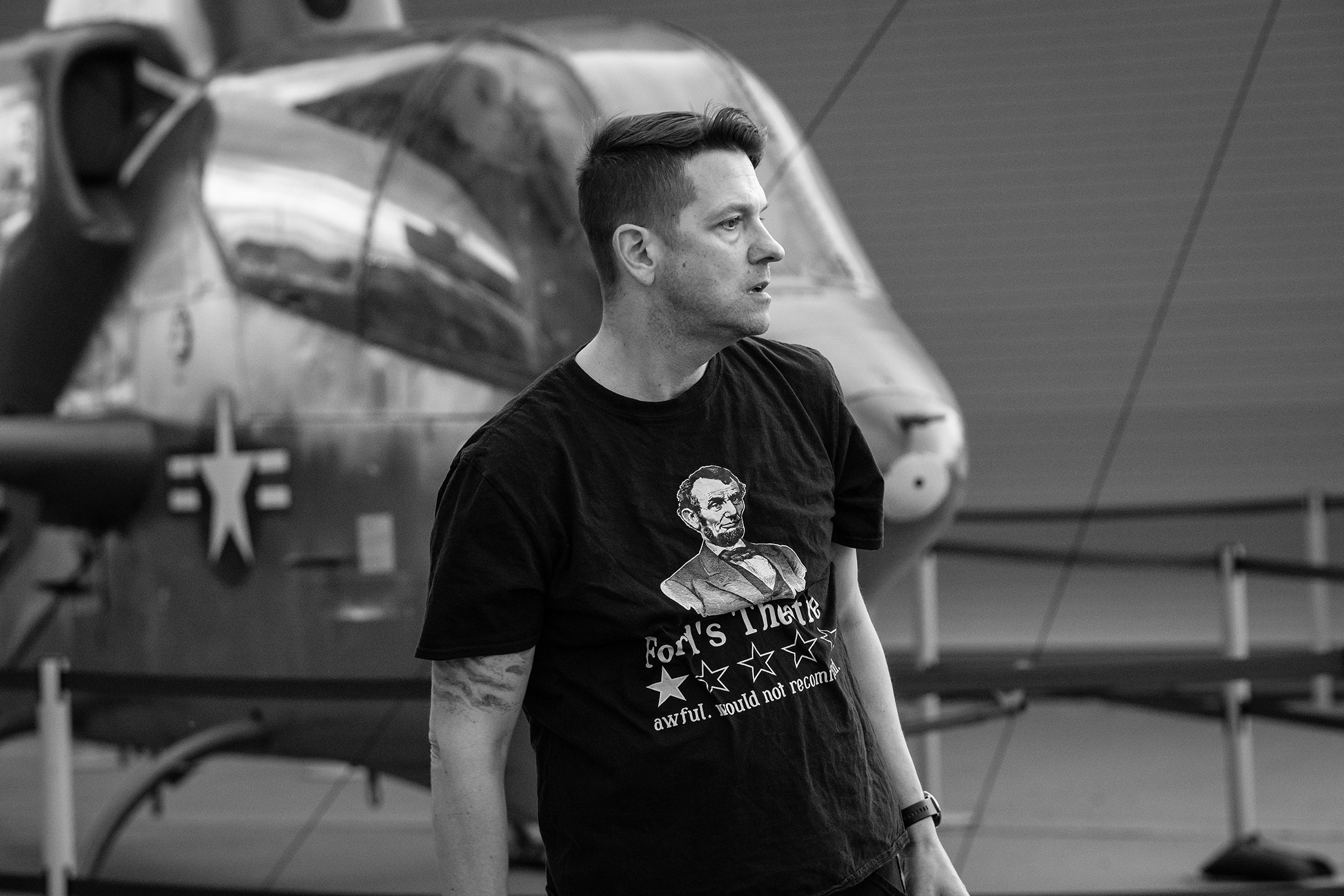 Black and white image of a white male with short dark hair wearing a t-shirt with an image of President Lincoln while looking to the right. There is a historic military plane in the background.