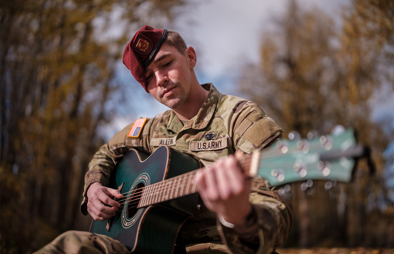 Young white man in a U.S. Army uniform sits outside playing a guitar.